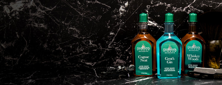 Three bottles of Clubman Pinaud aftershave products on a dark marble surface.