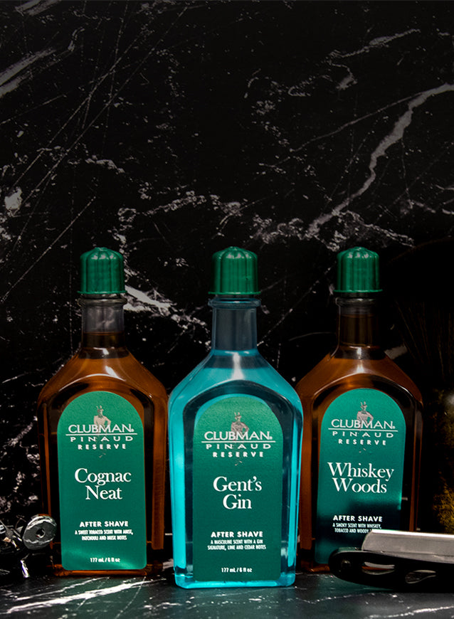 Three bottles of Clubman Pinaud aftershave products on a dark marble surface.