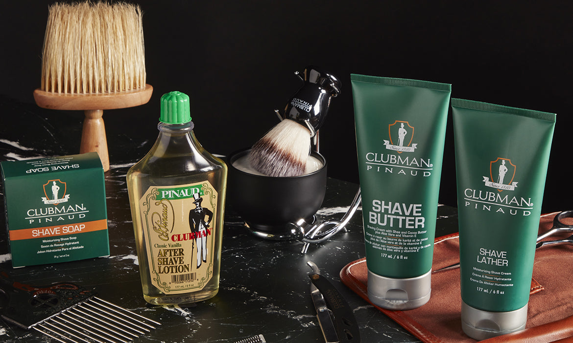 Clubman Pinaud shave soap, after shave lotion, shave butter, and shave lather displayed with a shaving brush and bowl on a black marble surface.