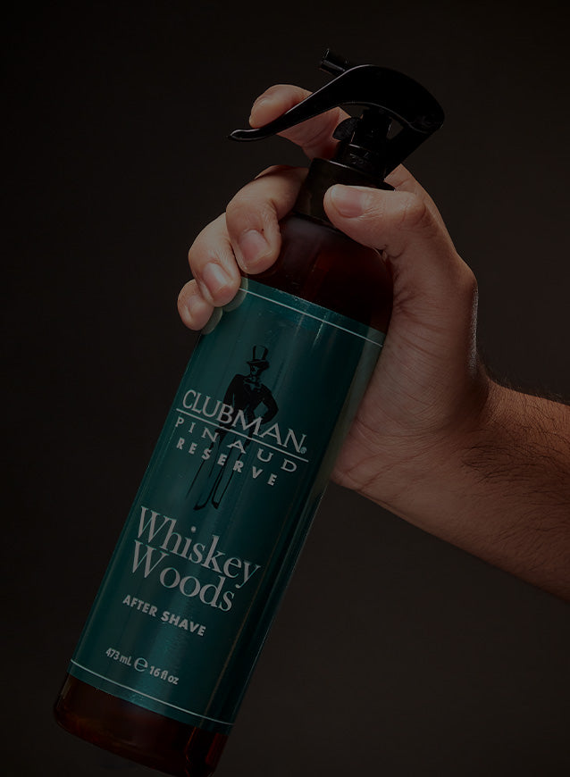 Hand holding a bottle of Clubman Pinaud Reserve Whiskey Woods After Shave against a dark background
