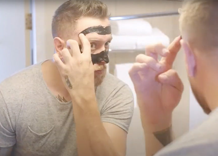 Man applying a black facial mask in front of a mirror, wearing a gray t-shirt.