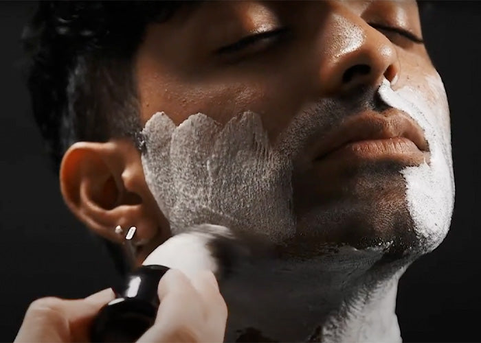 Man lathering shaving cream on his face with a black shaving brush, preparing for a clean shave.